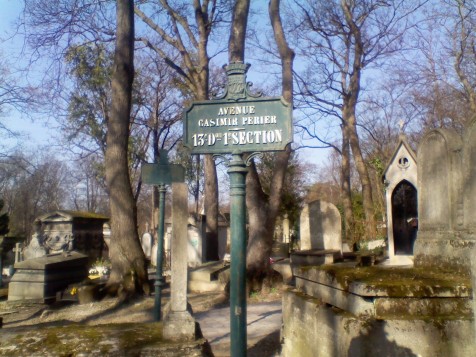 A dark green sign in front of some trees and monuments, with white letters reading 'AVENUE CASIMIR PERIER 13e Don 1e SECTION'