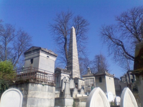 A cluster of white stone monuments, with a tall obelisk in the centre. In the background are several tall leafless trees