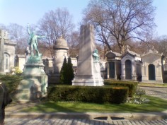 Two monuments with bronze statues: one white bier with a green statue of a man wearing an open jacket with one arm pointing upwards and one to the right, the other a tall white obelisk-like monument with a green bust of a bald man on its right-hand side