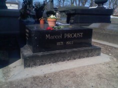 A black marble slab with the words 'Marcel PROUST 1871-1922' inscribed in gold letters. On top of the slab are a wilting red rose and two terracotta pots, one containing white flowers and one containing flowers of indistinguishable colour
