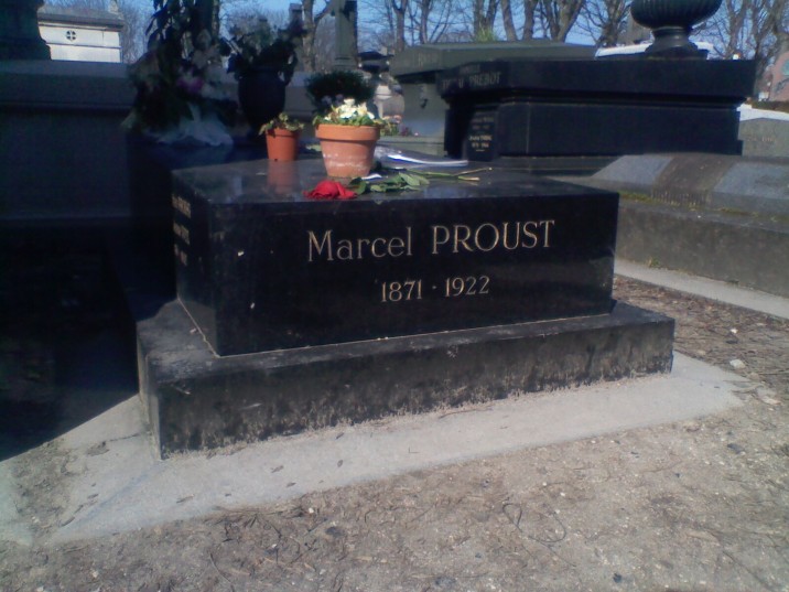 A black marble slab with the words 'Marcel PROUST 1871-1922' inscribed in gold letters. On top of the slab are a wilting red rose and two terracotta pots, one containing white flowers and one containing flowers of indistinguishable colour