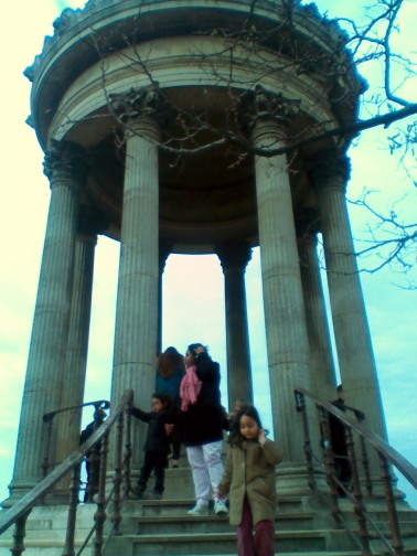 View from below of an ornate rotunda, with a circular roof supported by six columns in the classical style. On the steps leading up are a woman in a black coat and pink scarf, and two children