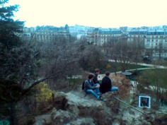 Three men sit on a rocky outcrop overlooking a park with many trees, mostly leafless. In the background many buildings are visible, including (silhoutted against the sky) the dome of the Basilique du Sacré-Cœur