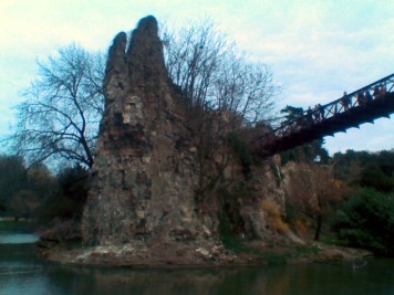 A tall, craggy rock promontory, covered in trees, rises almost vertically out of a green lake. To the right a bridge leads across to it