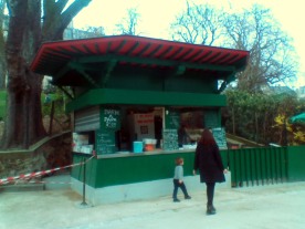 A green food and drinks stand with a flat red roof supported by green beams. Walking by in front are a child wearing a blue shirt and a woman in a black coat and leggings