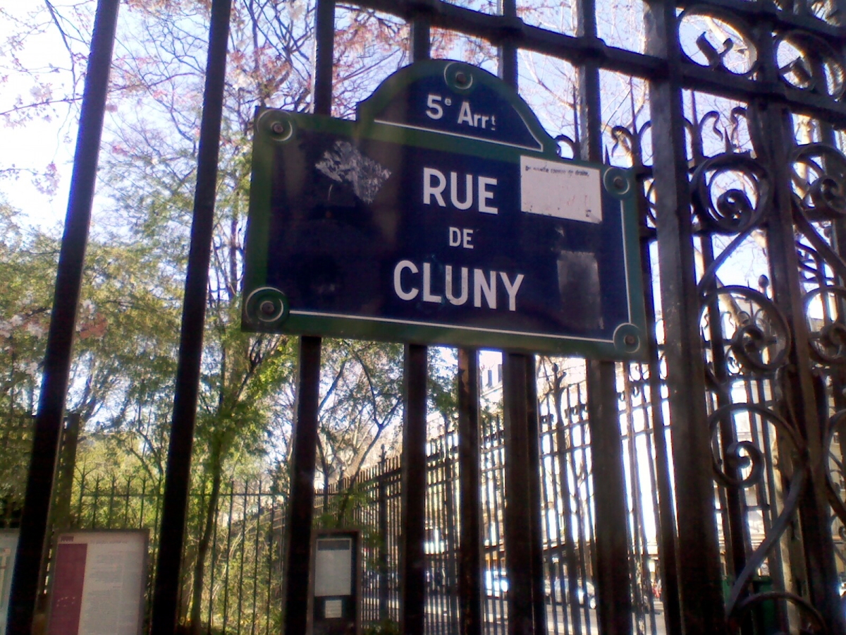 A blue and green sign reading '5e Arrt RUE DU CLUNY' is mounted on a black metal fence