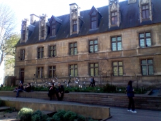 People stroll between garden beds in the kitchen garden of the Jardin du musée de Cluny. In the background is a long three-storey building with a dark blue roof and many windows