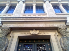 Top of a large, imposing set of dark green double doors. Enormous caryatids stand to either side; above, a stone inscription reads 'FACULTE DE MEDECINE'