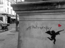 A statue plinth; drawn on it are a silhouette of a winged cherub holding a rifle, a small red heart, and the hashtag '#MTPhotography' (apart from the heart, b&w)