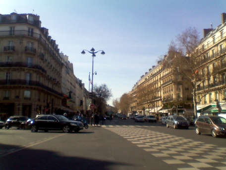 View down the Boulevard Saint-Germain in the afternoon; five- and six-storey buildings line either side. There are numerous pedestrians and small, mostly dark-coloured cars