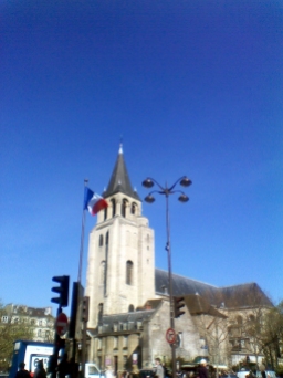 The steeple of the Église de Saint-Germain-des-Prés against a clear blue sky. In front are a flagpole bearing the tricolour of France and a tall lamppost with four lamps