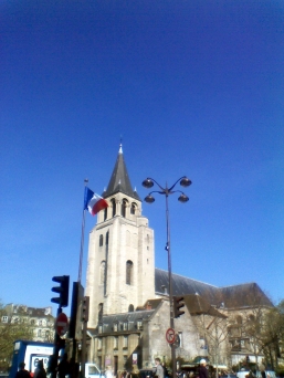 The steeple of the Église de Saint-Germain-des-Prés against a clear blue sky. In front are a flagpole bearing the tricolour of France and a tall lamppost with four lamps