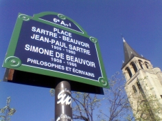 A blue and green sign reads '6e Arrt / PLACE SARTRE-BEAUVOIR / JEAN-PAUL SARTRE 1905-1980 / SIMONE DE BEAUVOIR 1908-1986 / PHILOSOPHES ET ÉCRIVAINS'. To the right of the sign is a tree with small new leaves and a tall church steeple