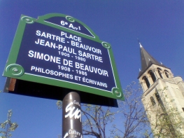 A blue and green sign reads '6e Arrt / PLACE SARTRE-BEAUVOIR / JEAN-PAUL SARTRE 1905-1980 / SIMONE DE BEAUVOIR 1908-1986 / PHILOSOPHES ET ÉCRIVAINS'. To the right of the sign is a tree with small new leaves and a tall church steeple