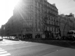 Bright sunlight shines over a road, with two pedestrian crossings visible to the left. A large, blocky multistorey building casts a long shadow over the road to the right (b&w)