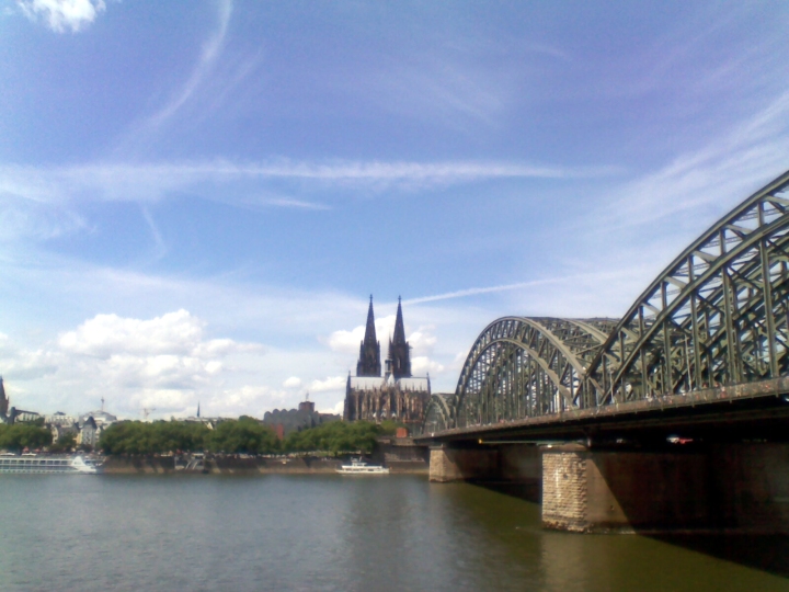 View of the Kölner Dom from the eastern bank of the Rhine. Leading to the cathedral is a large iron bridge mounted on stone pillars; one and a half of its long metal arches are visible. The greenish river water reflects the blue, partly cloudy sky above
