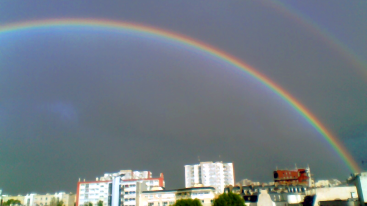 A large rainbow curves across an overcast sky. Below it, several apartment blocks and the leafy tops of trees are visible. Above it and to the right appears the fainter double rainbow.